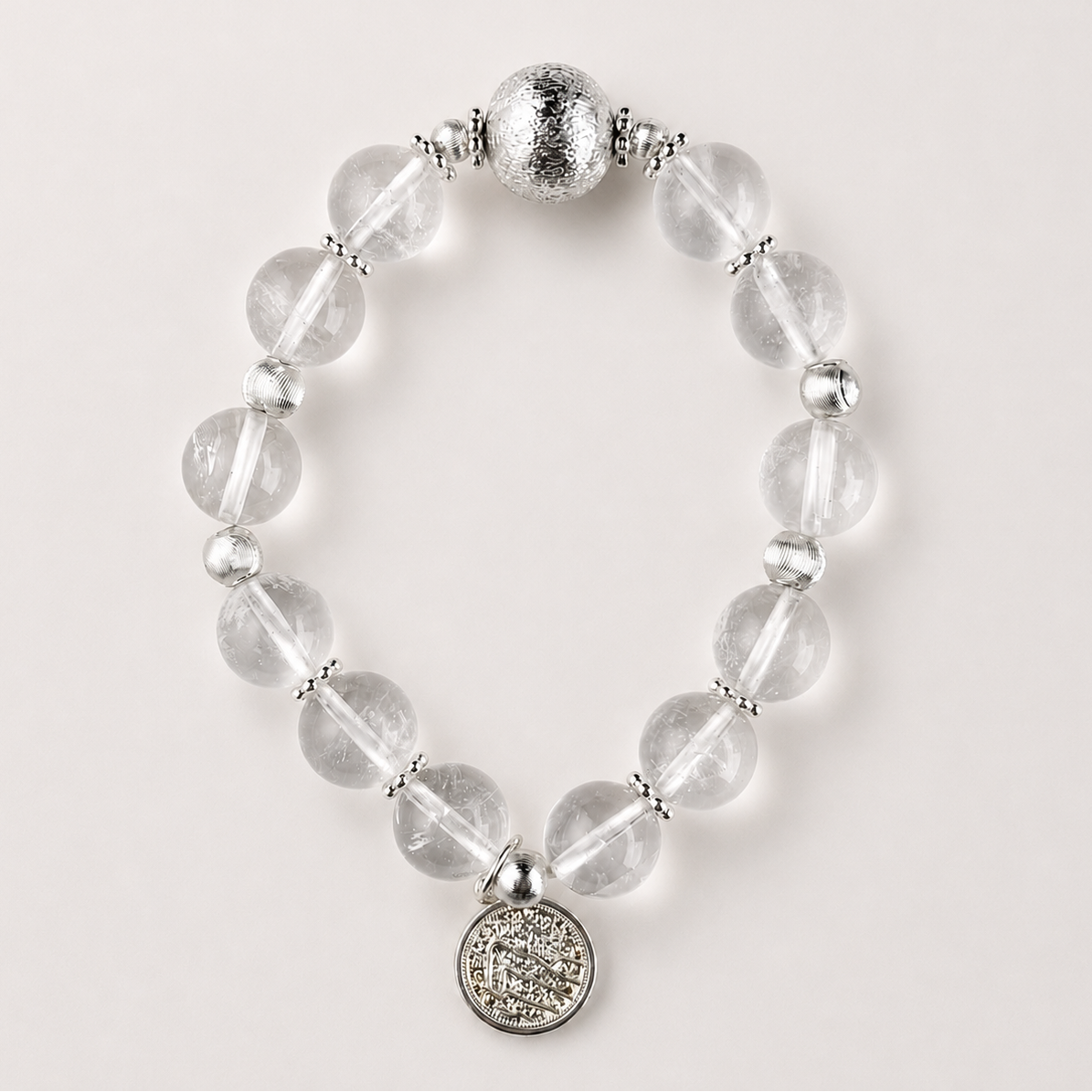 Clear quartz beaded bracelet with silver-toned accents and round coin charm on a soft light gray background.