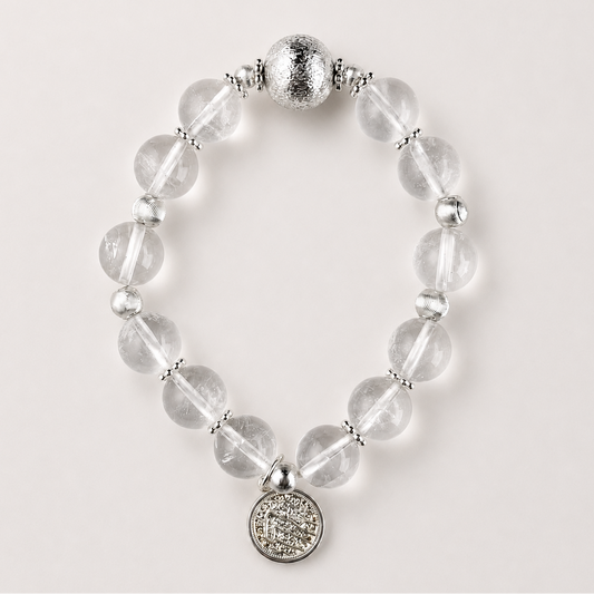 Clear quartz beaded bracelet with silver-toned accents and round coin charm on a soft light gray background.